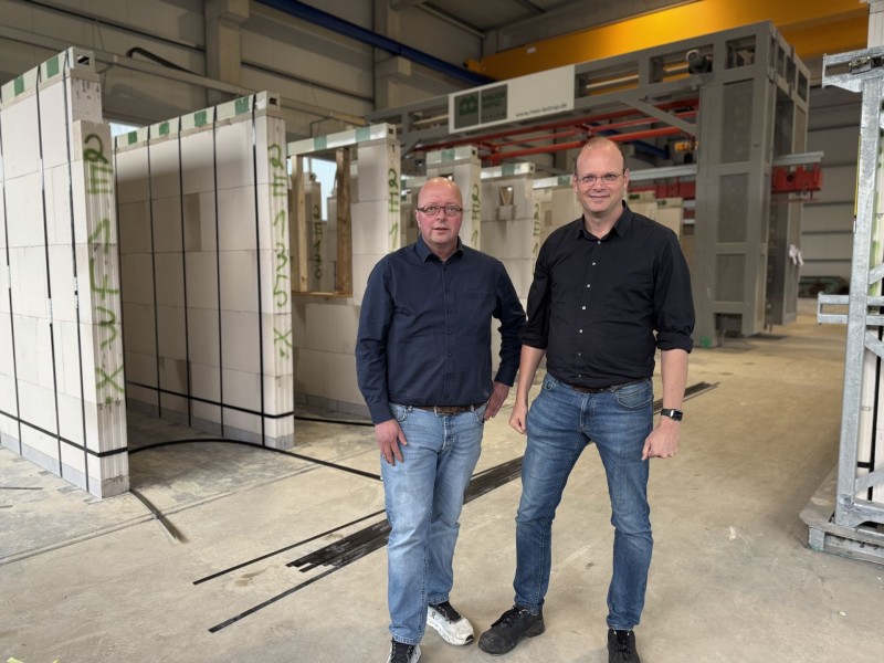 Zwei Männer stehen nebeneinander vor aufgereihten Betonwänden.