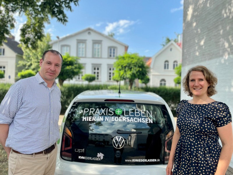 Eine Frau und ein Mann die vor einem weißen Gebäude rechts und links neben einem weißen Kleinwagen stehen