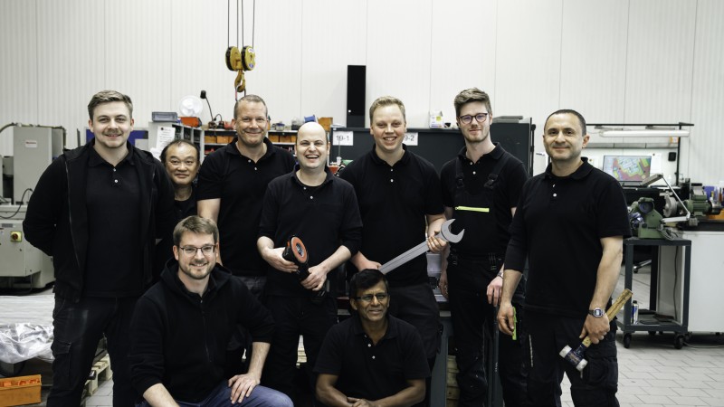 Team von Mitarbeitenden in schwarzer Arbeitskleidung posiert in einer Werkstattumgebung mit Werkzeugen und Maschinen.