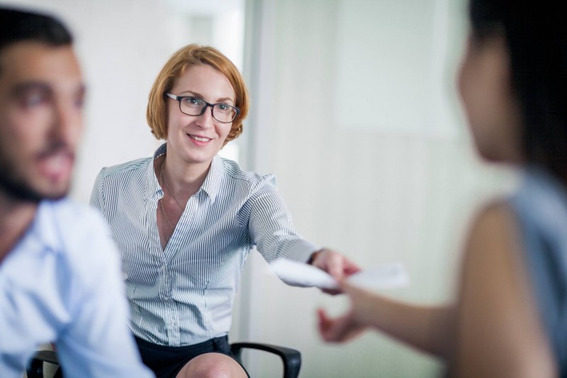 Frau mit kurzen Haaren und Brille übergibt ein Dokument an eine andere Person in einem Büro.